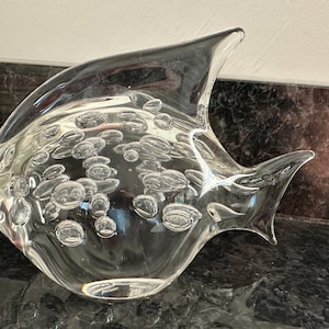 May include: A clear glass fish sculpture with internal bubbles. The fish has a curved body and fins, with a smooth, glossy finish. The bubbles are various sizes and shapes, adding visual interest. The sculpture is a decorative item.