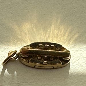 May include: A small, oval-shaped gold-toned metal charm featuring a horizontal bar across the centre and raised rectangular details. The charm includes a small circular jump ring attached to one side, designed for connecting to a chain or bracelet.