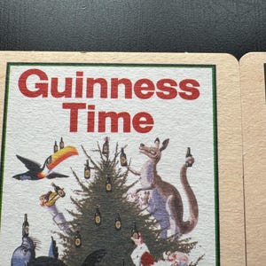 Puede incluir: Un posavasos vintage de Guinness Time con una ilustraci&oacute;n festiva. La imagen presenta un tuc&aacute;n, un canguro y otros animales alrededor de un &aacute;rbol de Navidad decorado con botellas de Guinness. El texto "Guinness Time" es rojo.
