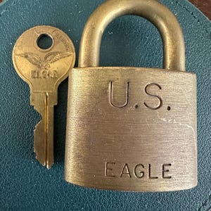 May include: A brass padlock with a U-shaped shackle and the text U.S. and EAGLE engraved on the front. Beside the padlock is a matching brass key featuring an eagle emblem and the word EAGLE with ILLINOIS stamped on the circular head.