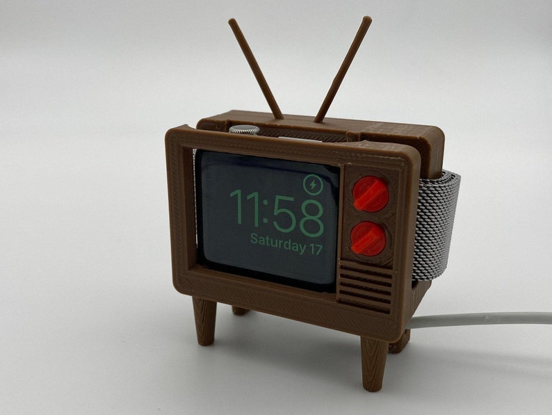 Puede incluir: Una r&eacute;plica de pl&aacute;stico marr&oacute;n de un televisor vintage con un Apple Watch cargando en su interior. La esfera del reloj muestra la hora 11:58 y la fecha s&aacute;bado 17. El televisor tiene una banda de metal plateado y dos botones rojos.