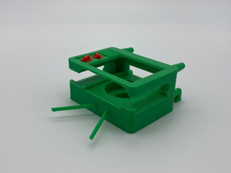 Puede incluir: Soporte de c&aacute;mara impreso en 3D de pl&aacute;stico verde con dos botones rojos en la parte superior.