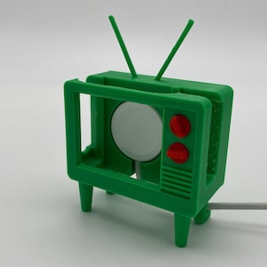 Puede incluir: Un soporte de televisi&oacute;n de pl&aacute;stico verde con un c&iacute;rculo blanco en el centro y dos botones rojos. El soporte tiene un cable blanco.