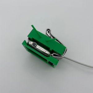 Puede incluir: Un soporte de impresi&oacute;n 3D verde para cargar un Apple Watch. El reloj est&aacute; en el soporte con una banda de metal. El soporte tiene dos peque&ntilde;as puntas en la parte superior.