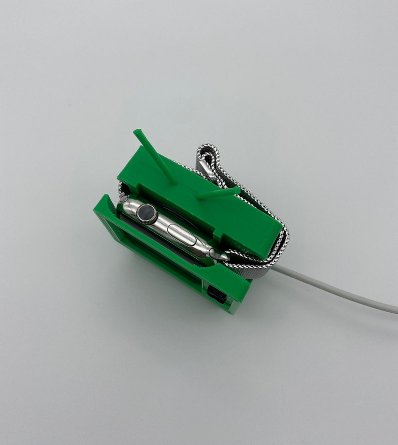 Puede incluir: Un soporte de impresi&oacute;n 3D verde para cargar un Apple Watch. El reloj est&aacute; en el soporte con una banda de metal. El soporte tiene dos peque&ntilde;as puntas en la parte superior.