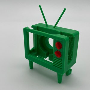 Puede incluir: Un modelo de televisi&oacute;n vintage de pl&aacute;stico verde con botones rojos y antenas. La televisi&oacute;n no tiene pantalla y tiene un interior hueco.