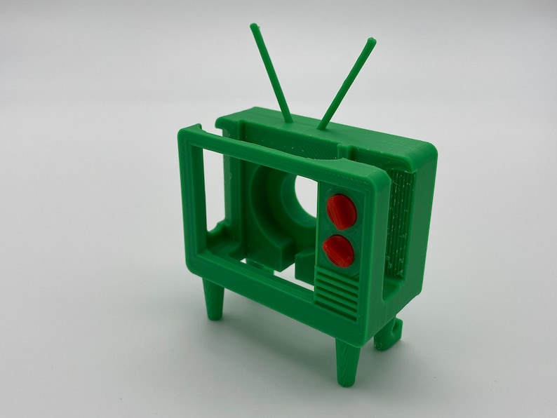 Puede incluir: Un modelo de televisi&oacute;n vintage de pl&aacute;stico verde con botones rojos y antenas. La televisi&oacute;n no tiene pantalla y tiene un interior hueco.