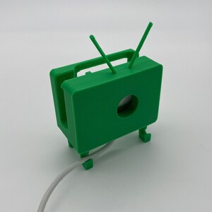 Puede incluir: Un modelo de televisi&oacute;n retro de color verde impreso en 3D con antenas. La televisi&oacute;n tiene un agujero en la parte delantera y un cable blanco est&aacute; conectado a la parte inferior.