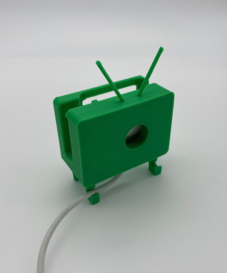 Puede incluir: Un modelo de televisi&oacute;n retro de color verde impreso en 3D con antenas. La televisi&oacute;n tiene un agujero en la parte delantera y un cable blanco est&aacute; conectado a la parte inferior.