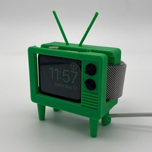 Puede incluir: Una caja de reloj de pl&aacute;stico verde con forma de televisor vintage con una correa de reloj a rayas blancas y negras. La esfera del reloj muestra la hora 11:57 y la fecha s&aacute;bado 17.