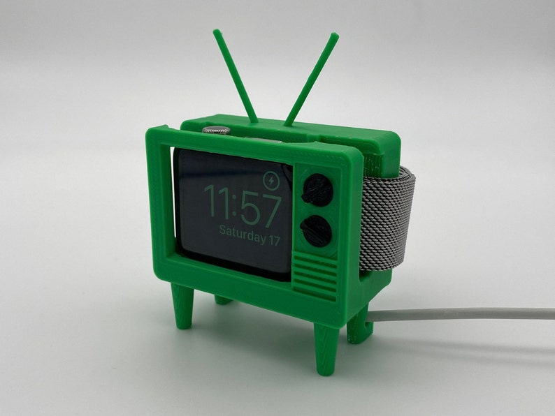 Puede incluir: Una caja de reloj de pl&aacute;stico verde con forma de televisor vintage con una correa de reloj a rayas blancas y negras. La esfera del reloj muestra la hora 11:57 y la fecha s&aacute;bado 17.