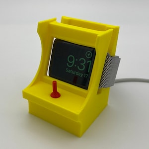May include: A yellow 3D printed stand shaped like an arcade game, with a red joystick, designed to hold and charge an Apple Watch. The watch face shows the time 9:31 and the date Saturday 17.