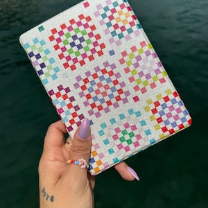 Pode incluir: Uma capa branca para leitor de e-books com um padrão xadrez colorido. A capa tem um acabamento brilhante e está sendo segurada em uma mão que está usando um anel de contas.