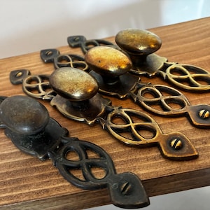 May include: Four antique brass drawer pulls with oval knobs and decorative backplates. The pulls are in varying shades of gold and bronze, with intricate openwork designs. They are mounted on a dark wooden surface.