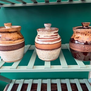 May include: Three ceramic jars with lids, each featuring a distinct glaze pattern. The left jar has a tan and brown finish, the centre jar displays horizontal brown and cream stripes, and the right jar features a deep brown base with a tan top.