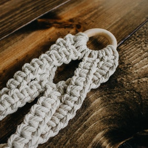 May include: A beige macrame plant hanger with a wooden ring at the top. The hanger is made of thick, knotted rope and has a unique, woven design.