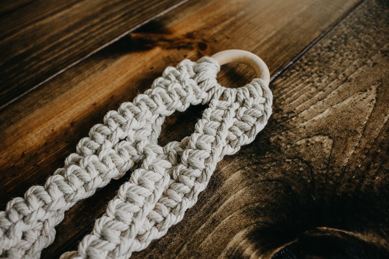 May include: A beige macrame plant hanger with a wooden ring at the top. The hanger is made of thick, knotted rope and has a unique, woven design.