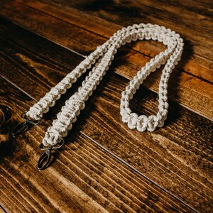 May include: A white cotton camera strap with two metal clips. The strap is made of a braided rope and is lying on a wooden surface.