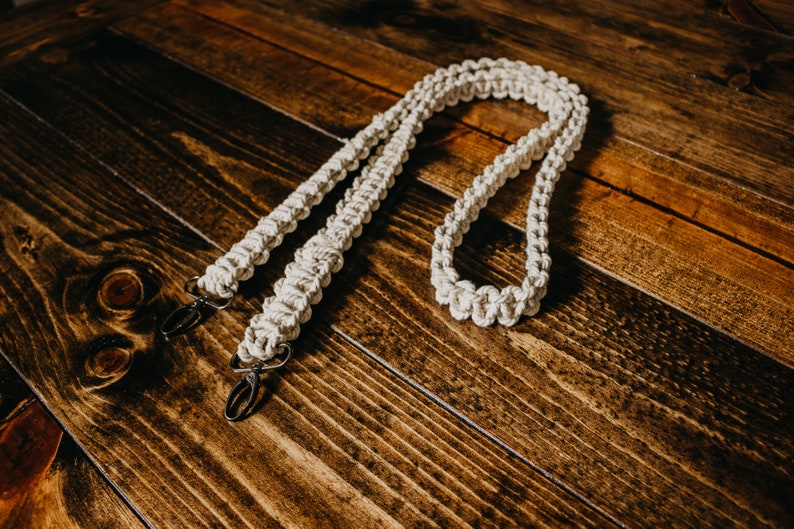 May include: A white cotton camera strap with two metal clips. The strap is made of a braided rope and is lying on a wooden surface.