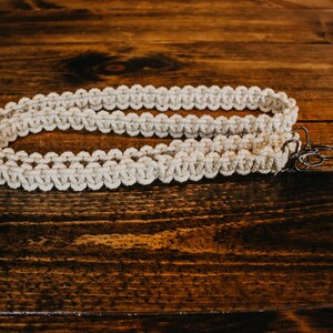 May include: A white macrame strap with two silver metal clasps. The strap is lying on a wooden surface.