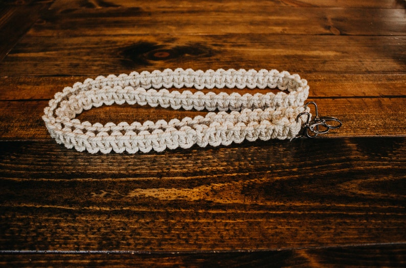 May include: A white macrame strap with two silver metal clasps. The strap is lying on a wooden surface.