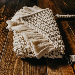 May include: A cream-colored macrame bag with a fringe detail and a long strap. The bag is made of natural fibers and has a bohemian style.