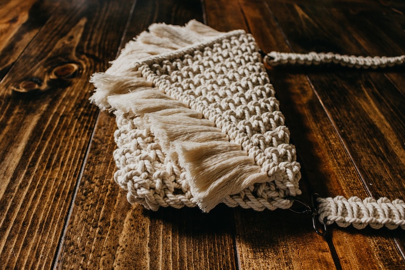 May include: A cream-colored macrame bag with a fringe detail and a long strap. The bag is made of natural fibers and has a bohemian style.