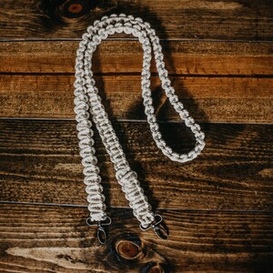 May include: A white macrame camera strap with two metal clasps. The strap is made of a thick, braided cord and is designed to be worn around the neck.