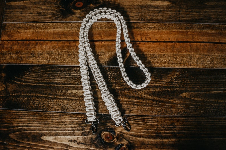 May include: A white macrame camera strap with two metal clasps. The strap is made of a thick, braided cord and is designed to be worn around the neck.