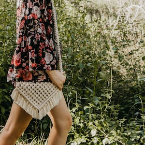 May include: A woman wearing a floral print blouse and a white crocheted bag with fringe. The bag is worn over the shoulder and is hanging down the side of the woman's body. The woman is standing in a grassy area.