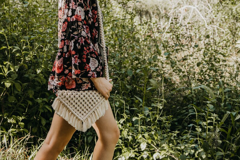 May include: A woman wearing a floral print blouse and a white crocheted bag with fringe. The bag is worn over the shoulder and is hanging down the side of the woman's body. The woman is standing in a grassy area.