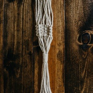 May include: A white macrame plant hanger with a tassel hanging against a wooden background.