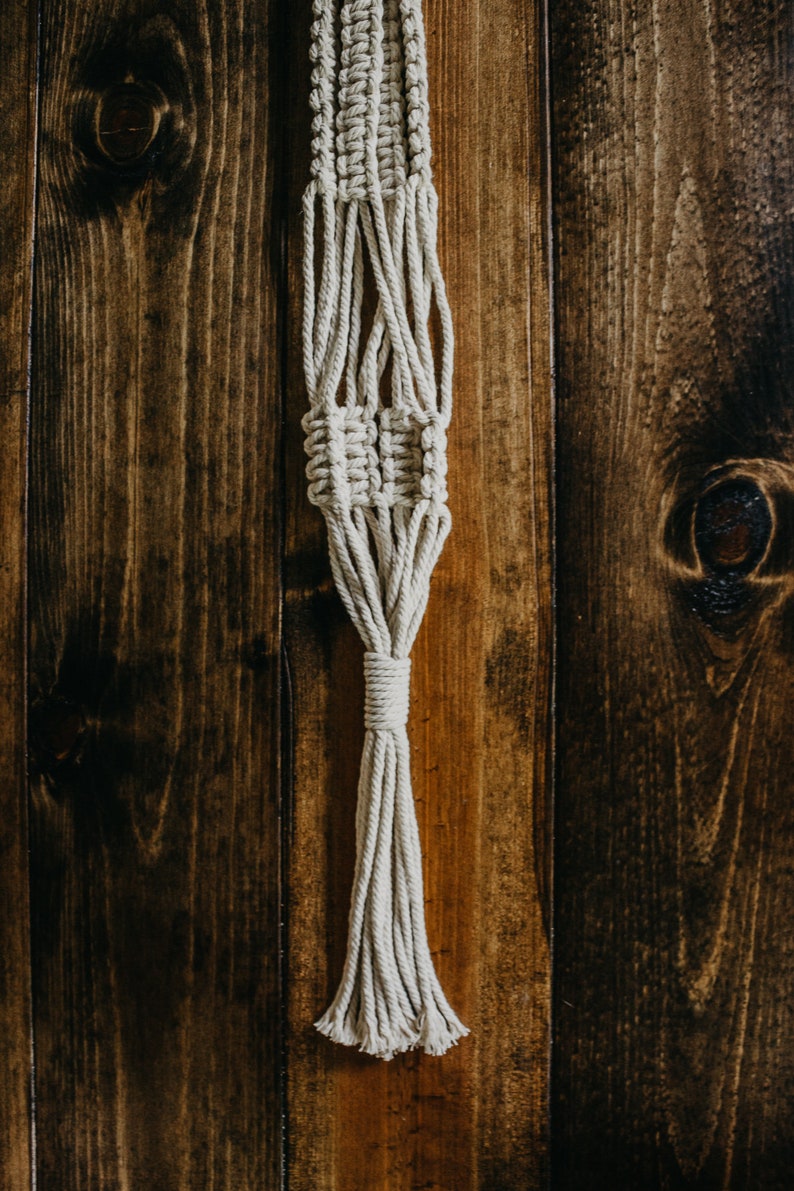 May include: A white macrame plant hanger with a tassel hanging against a wooden background.