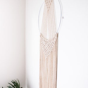 Op de afbeelding: Een beige macram&eacute; wandkleed met een cirkelvormig metalen frame. Het macram&eacute; heeft een geweven ontwerp aan de bovenkant en lange, vloeiende strengen. De wanddecoratie hangt tegen een witte muur en voegt een vleugje bohemien stijl toe.