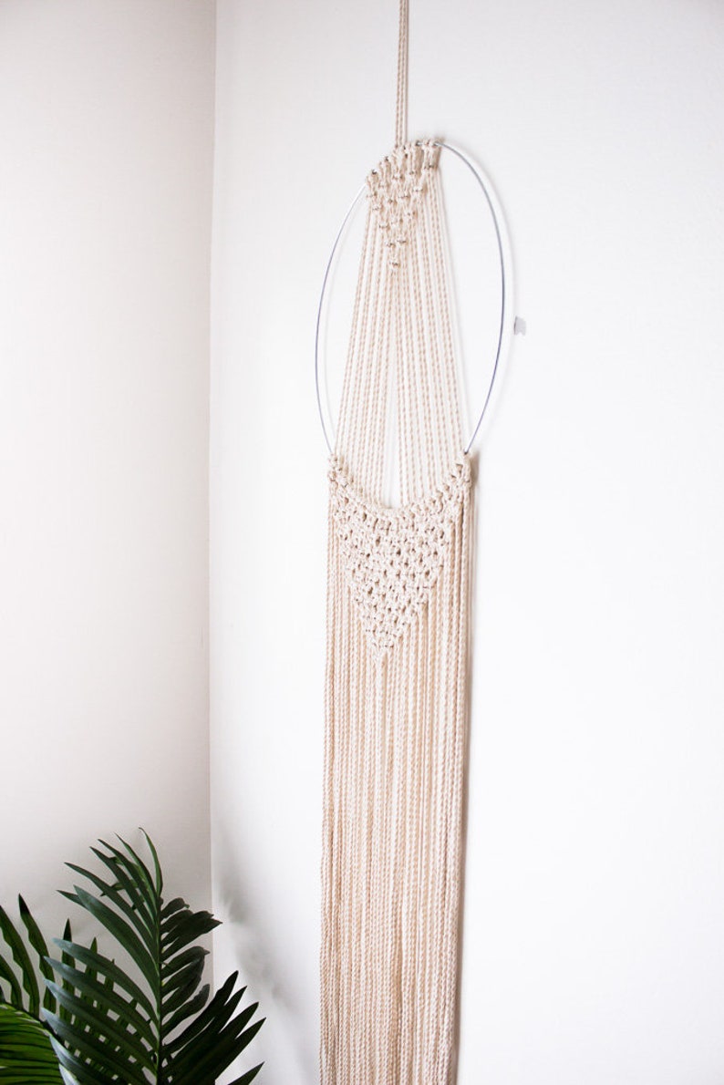 Op de afbeelding: Een beige macram&eacute; wandkleed met een cirkelvormig metalen frame. Het macram&eacute; heeft een geweven ontwerp aan de bovenkant en lange, vloeiende strengen. De wanddecoratie hangt tegen een witte muur en voegt een vleugje bohemien stijl toe.