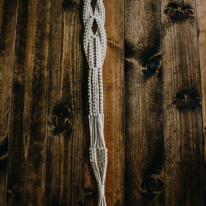 May include: A macrame plant hanger with a wooden ring at the top. The hanger is made of natural coloured cord and has a diamond pattern. It is hanging against a dark wood background.