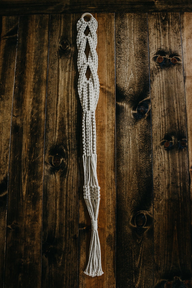 May include: A macrame plant hanger with a wooden ring at the top. The hanger is made of natural coloured cord and has a diamond pattern. It is hanging against a dark wood background.