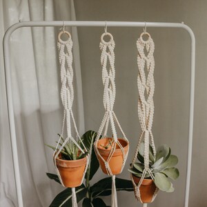 May include: Three macrame plant hangers with wooden rings hang from a white metal rack. Each hanger holds a terracotta pot with a green plant.