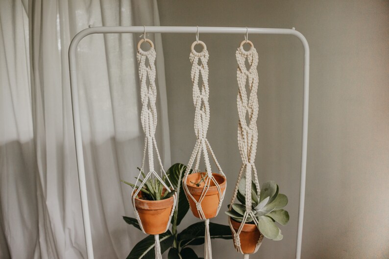 May include: Three macrame plant hangers with wooden rings hang from a white metal rack. Each hanger holds a terracotta pot with a green plant.