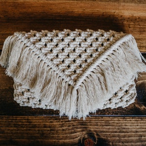 May include: A beige crocheted clutch with fringe detailing. The clutch is made with a macrame knot pattern and has a triangular shape.