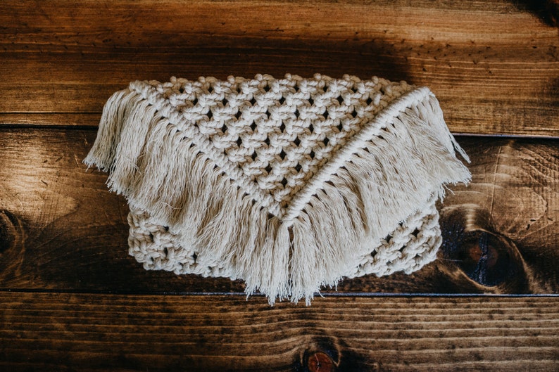 May include: A beige crocheted clutch with fringe detailing. The clutch is made with a macrame knot pattern and has a triangular shape.