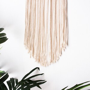 Op de afbeelding: Cr&egrave;mekleurige macram&eacute; wandhanger met een getextureerde, gefranjerde onderkant. De wanddecoratie wordt gepresenteerd tegen een witte achtergrond, met groene bladplanten op de voorgrond. Dit handgemaakte stuk voegt een bohemien touch toe.