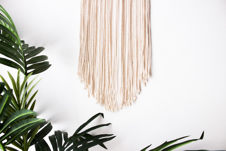 Op de afbeelding: Cr&egrave;mekleurige macram&eacute; wandhanger met een getextureerde, gefranjerde onderkant. De wanddecoratie wordt gepresenteerd tegen een witte achtergrond, met groene bladplanten op de voorgrond. Dit handgemaakte stuk voegt een bohemien touch toe.