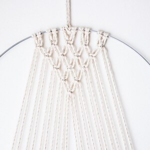 Op de afbeelding: Een macram&eacute; wandkleed met een geometrisch ontwerp. Het stuk heeft een zilveren metalen ring en off-white katoenen koord. Het geknoopte patroon vormt een reeks ruitvormen, met lange strengen koord eronder.