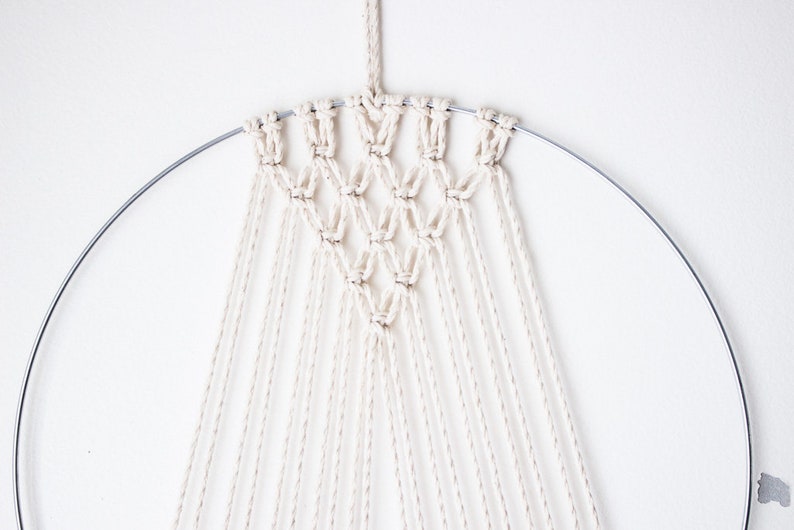 Op de afbeelding: Een macram&eacute; wandkleed met een geometrisch ontwerp. Het stuk heeft een zilveren metalen ring en off-white katoenen koord. Het geknoopte patroon vormt een reeks ruitvormen, met lange strengen koord eronder.