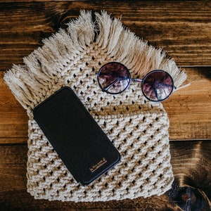 May include: A black phone case with a gold logo sits on a cream-colored crocheted bag with fringe. Round sunglasses with dark lenses are on top of the bag.