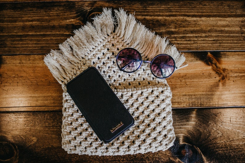 May include: A black phone case with a gold logo sits on a cream-colored crocheted bag with fringe. Round sunglasses with dark lenses are on top of the bag.
