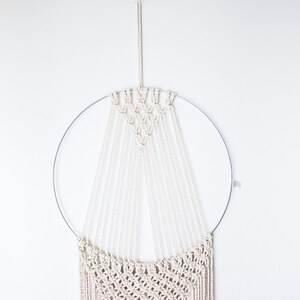 Op de afbeelding: Een macram&eacute; wandkleed met een cr&egrave;mekleurig ontwerp, opgehangen aan een zilveren metalen ring. Het ingewikkelde geweven patroon omvat een geometrisch bovendeel en vloeiende franjes, waardoor een bohemien esthetiek ontstaat. De wanddecoratie staat tegen een witte achtergrond.