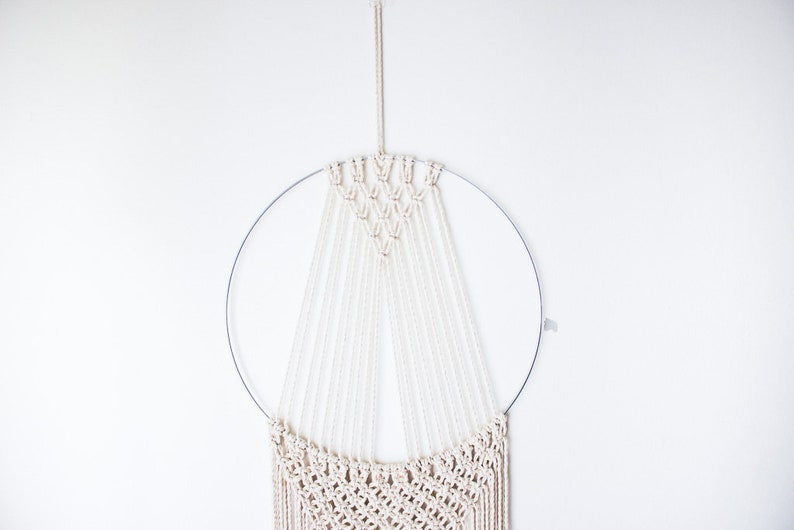 Op de afbeelding: Een macram&eacute; wandkleed met een cr&egrave;mekleurig ontwerp, opgehangen aan een zilveren metalen ring. Het ingewikkelde geweven patroon omvat een geometrisch bovendeel en vloeiende franjes, waardoor een bohemien esthetiek ontstaat. De wanddecoratie staat tegen een witte achtergrond.