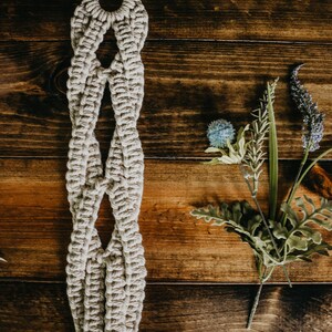 May include: A beige macrame plant hanger with a wooden ring at the top. The hanger is hanging against a dark brown wooden background.